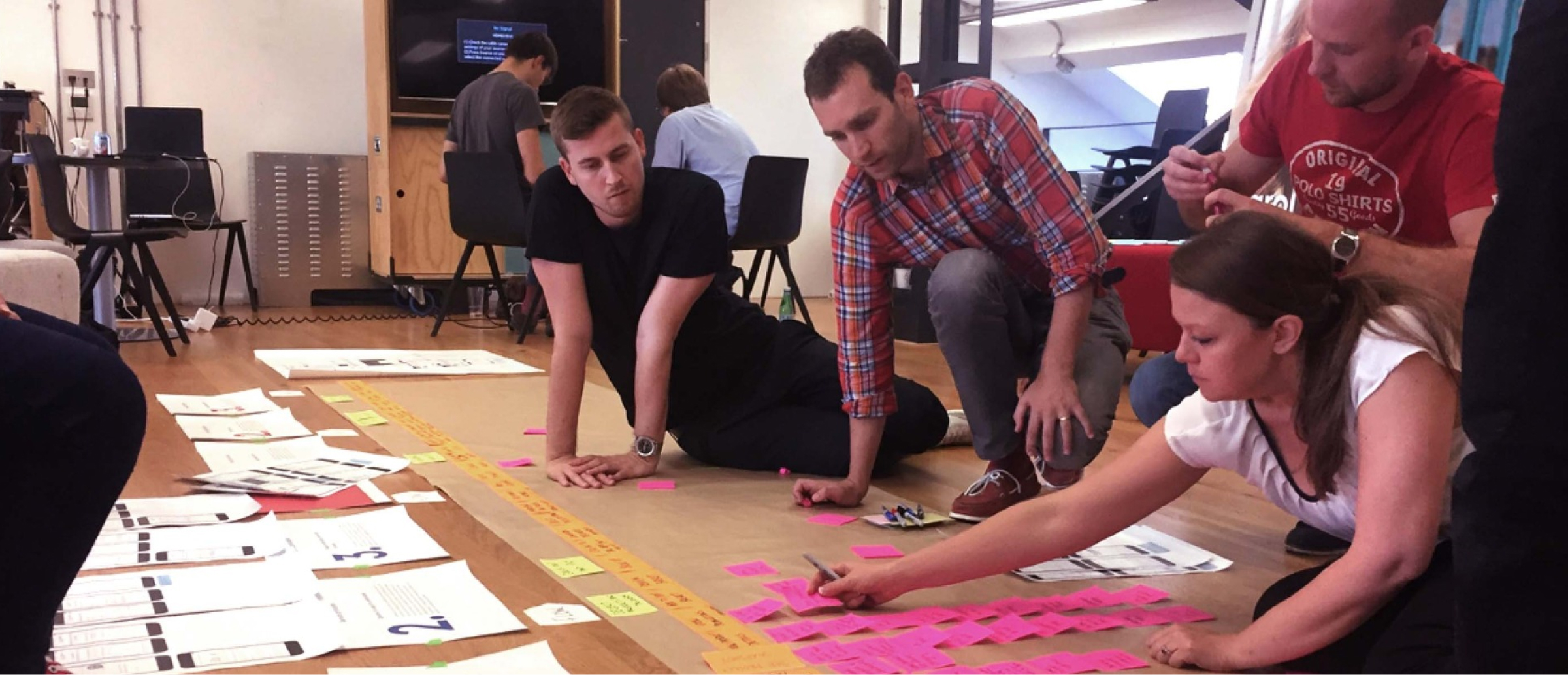 A group of people collaborating during a workshop, sitting on the floor and organizing notes on large sheets of paper. One person is writing on pink sticky notes, while others are reviewing documents spread out on the floor. The setting emphasizes teamwork and creative brainstorming