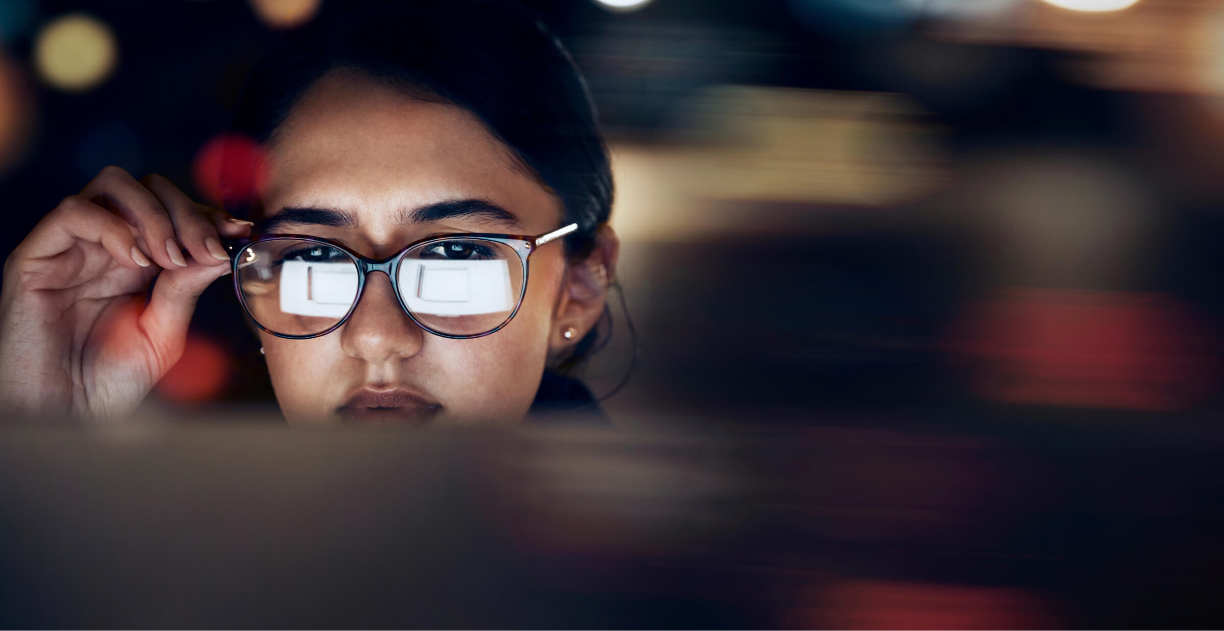A close-up of a woman wearing glasses, intensely focused on a screen, with the reflection of the screen visible in her lenses. The background is blurred, highlighting her concentration and the digital nature of her work.
