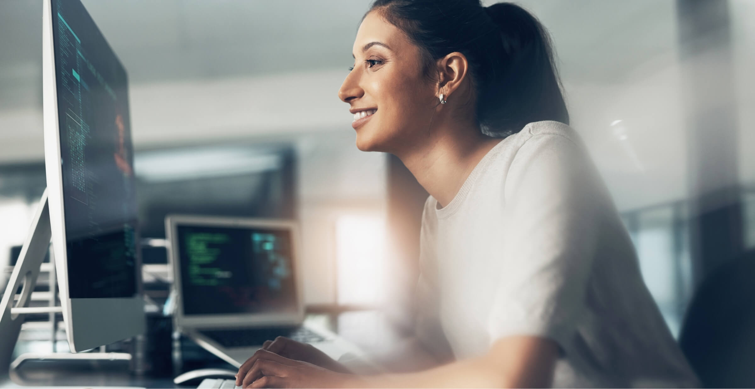 A woman smiling while working at a computer in a modern office. She is focused on the screen, which displays lines of code, with a second monitor visible in the background. The image conveys a positive work environment and engagement in programming or cybersecurity tasks.