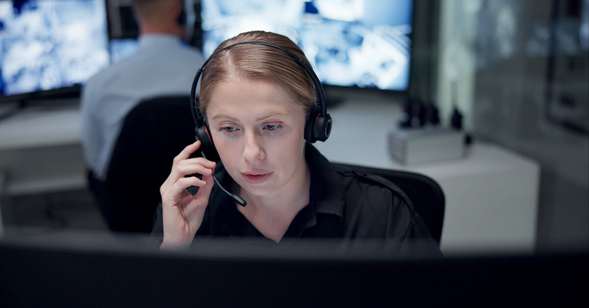 A focused woman wearing a headset works at a computer in a monitoring control room with screens in the background.