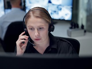A focused woman wearing a headset works at a computer in a monitoring control room with screens in the background.