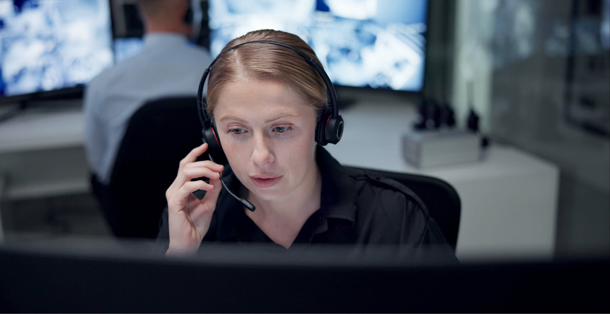 A focused woman wearing a headset works at a computer in a monitoring control room with screens in the background.