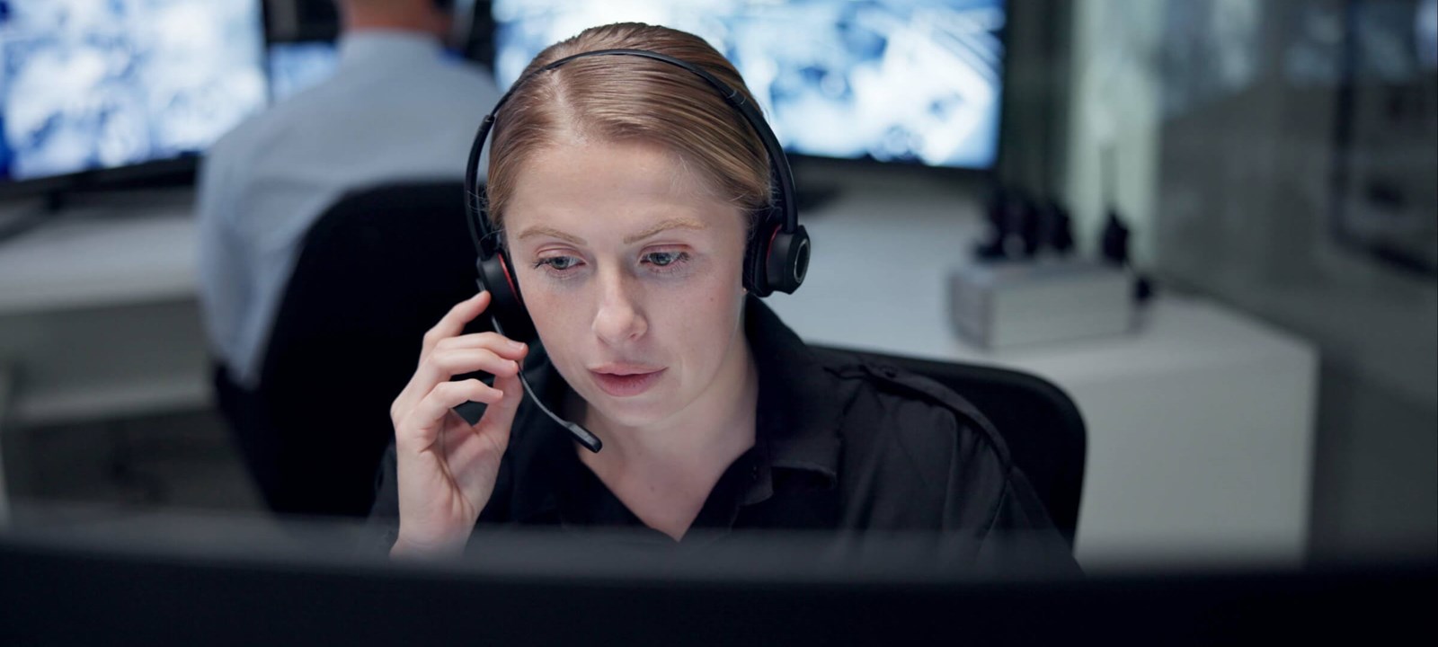 A focused woman wearing a headset works at a computer in a monitoring control room with screens in the background.