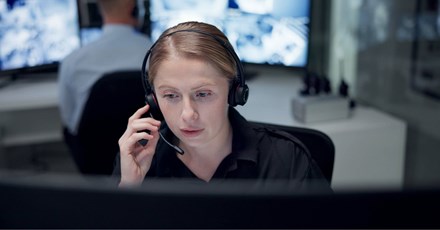 A focused woman wearing a headset works at a computer in a monitoring control room with screens in the background.