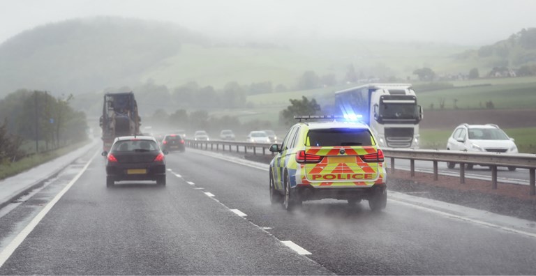 A police car with flashing lights drives on a wet highway in rainy weather, surrounded by other vehicles.