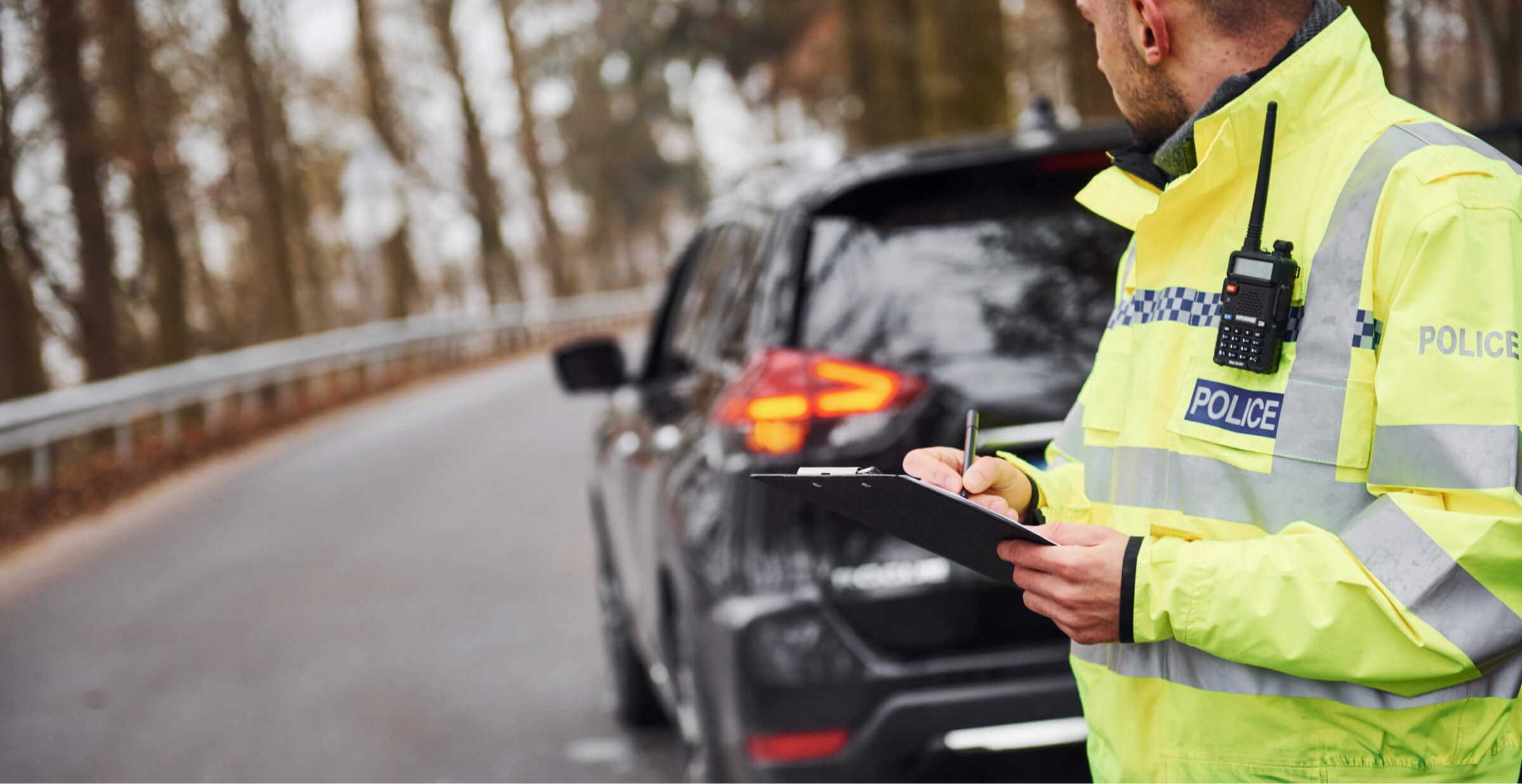 Police officer in high-visibility jacket writing a report near a black car on a forest road, ensuring road safety and law enforcement.