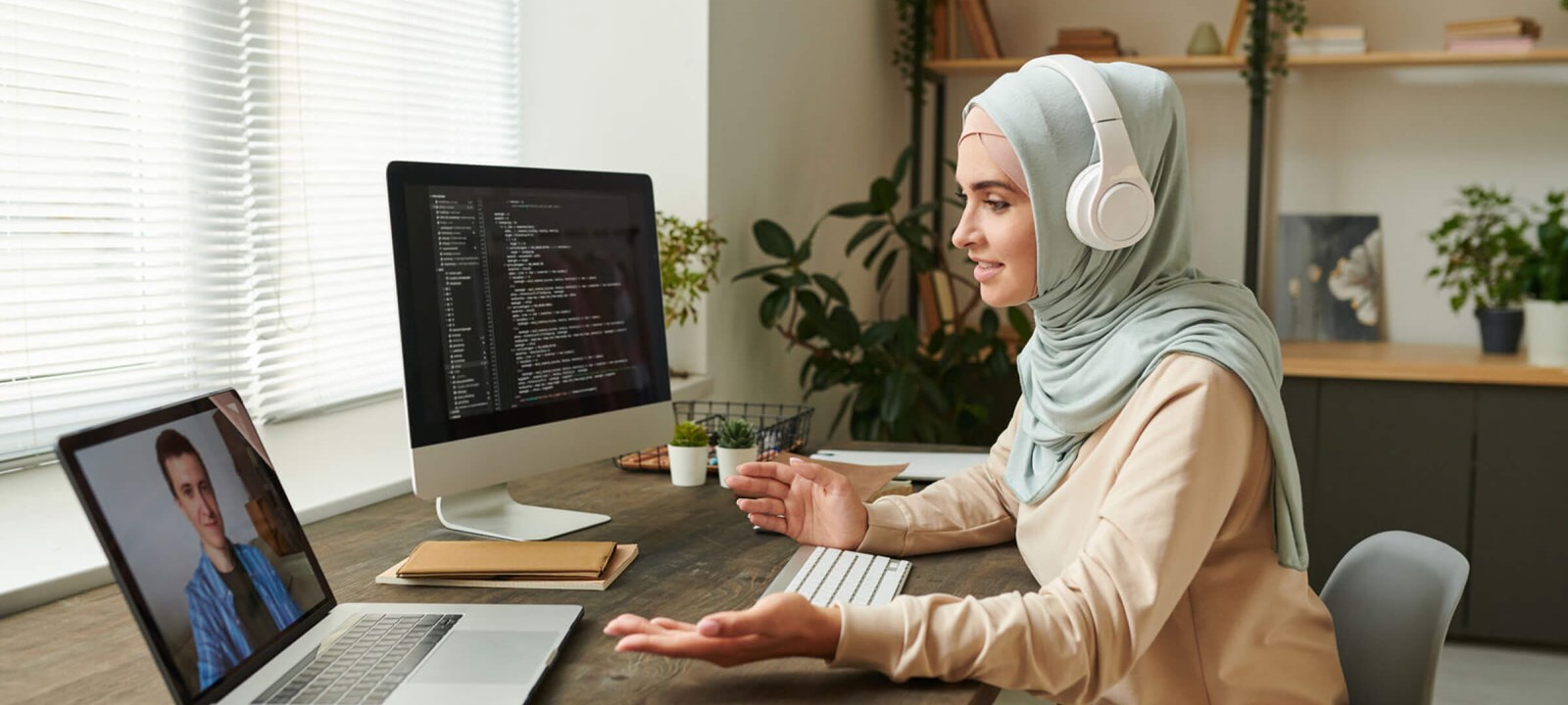A woman wearing a hijab and headphones working from home, participating in a video call on her laptop while coding on a desktop computer. She is seated at a desk in a modern home office with plants and shelves in the background, engaged in a remote work setting.