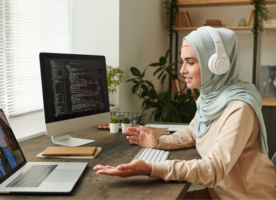 A woman wearing a hijab and headphones working from home, participating in a video call on her laptop while coding on a desktop computer. She is seated at a desk in a modern home office with plants and shelves in the background, engaged in a remote work setting.
