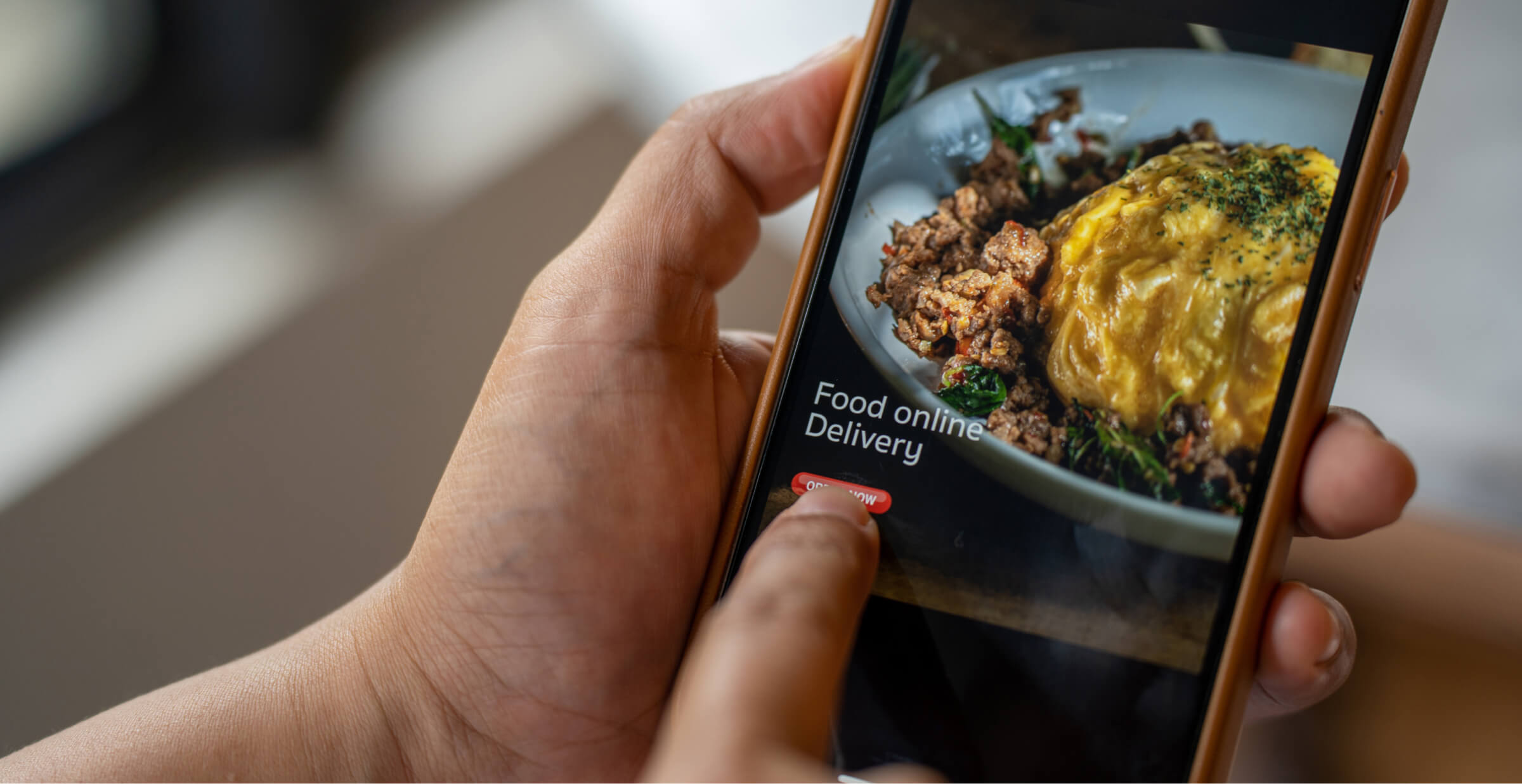 A close-up of a person using a smartphone to order food online. The screen shows a dish of food with the text 'Food online delivery' and a red 'Order Now' button, highlighting the convenience of mobile food ordering