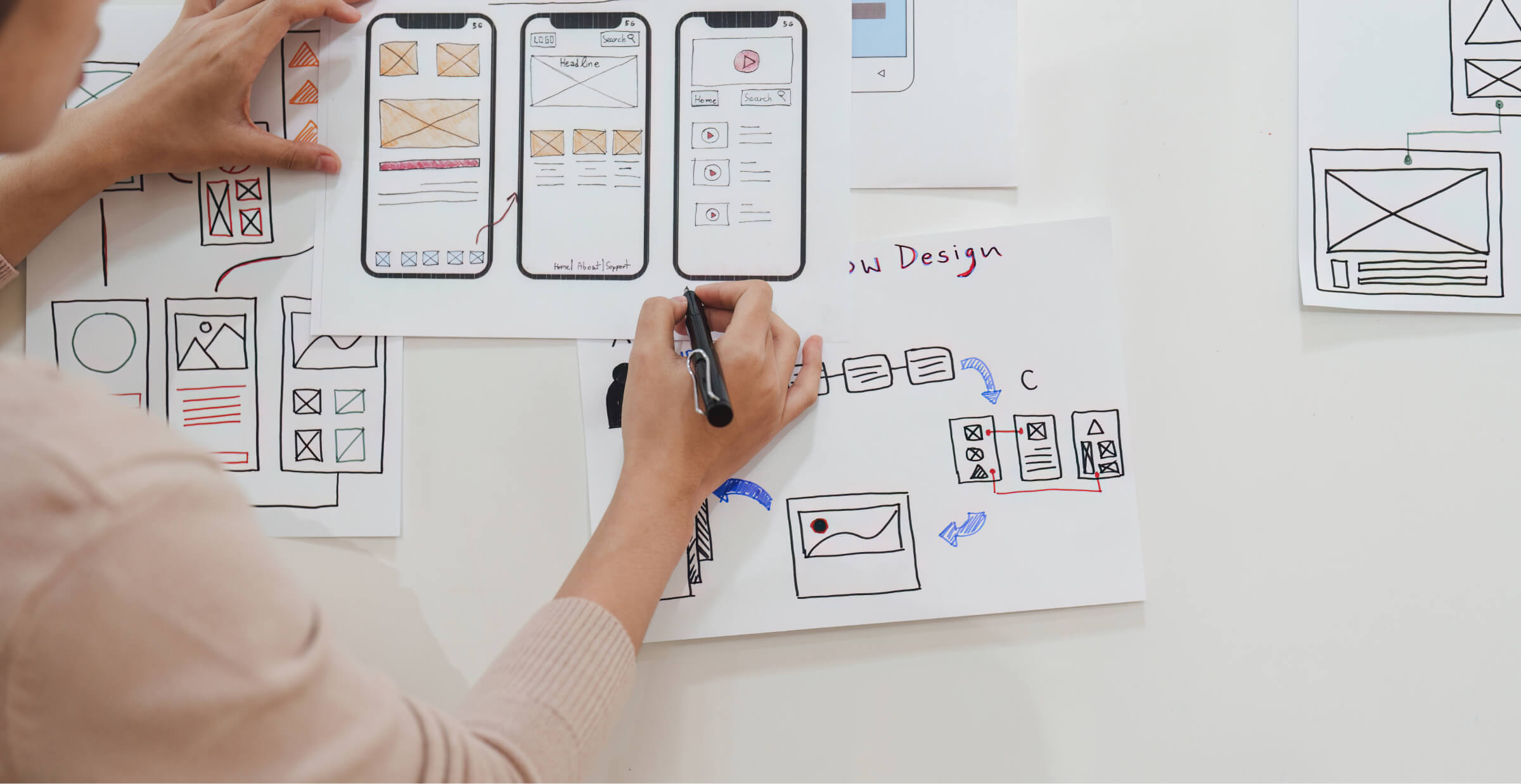 A designer working on app wireframes, sketching out the flow and layout of a mobile application on paper. Various screens and interface designs are pinned to the wall, illustrating the app's structure and user experience. The designer is actively drawing connections between different screens.