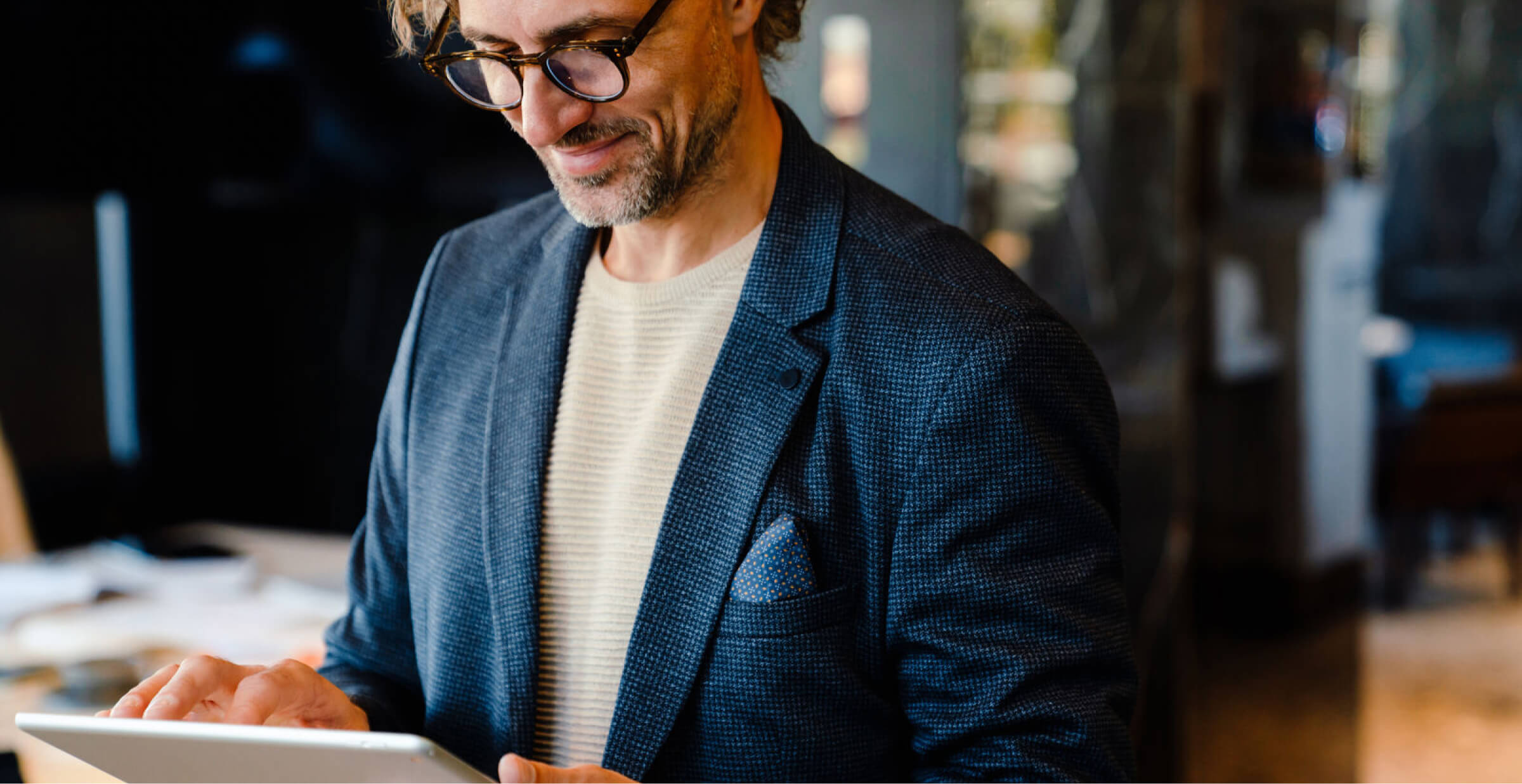 A man with glasses and a light beard, wearing a blue blazer and a light sweater, is using a tablet in a modern office environment. He is smiling and focused on the device. The background is softly blurred, suggesting a professional workspace, with desks and papers visible. The atmosphere appears calm and productive.