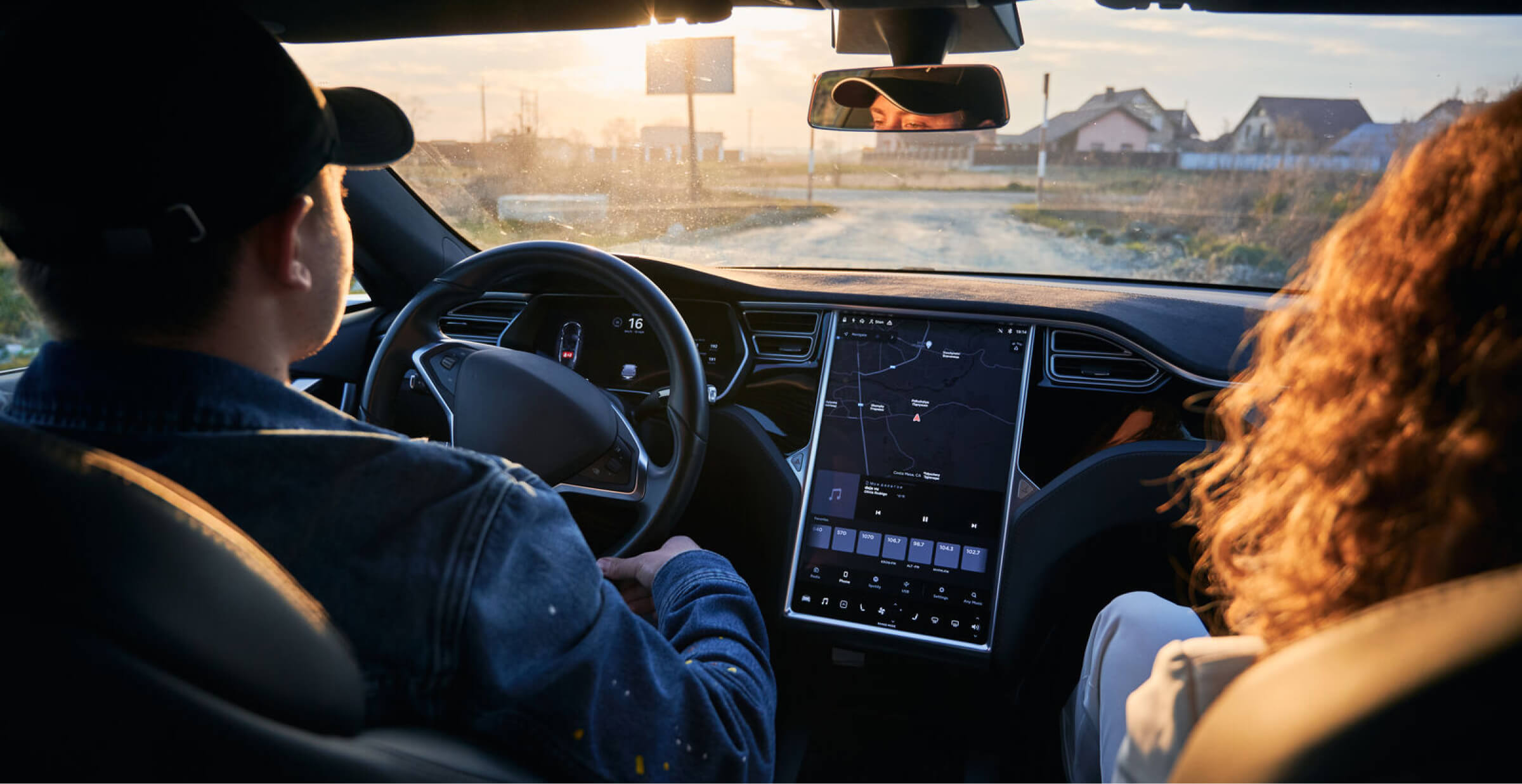 A view from the inside of a modern car showing two people in the front seats, one driving and the other as a passenger. The car has a large digital display in the center console displaying a map and controls, indicating advanced technology in use. The scene is set during sunset, giving a warm glow to the interior, with houses visible through the windshield.