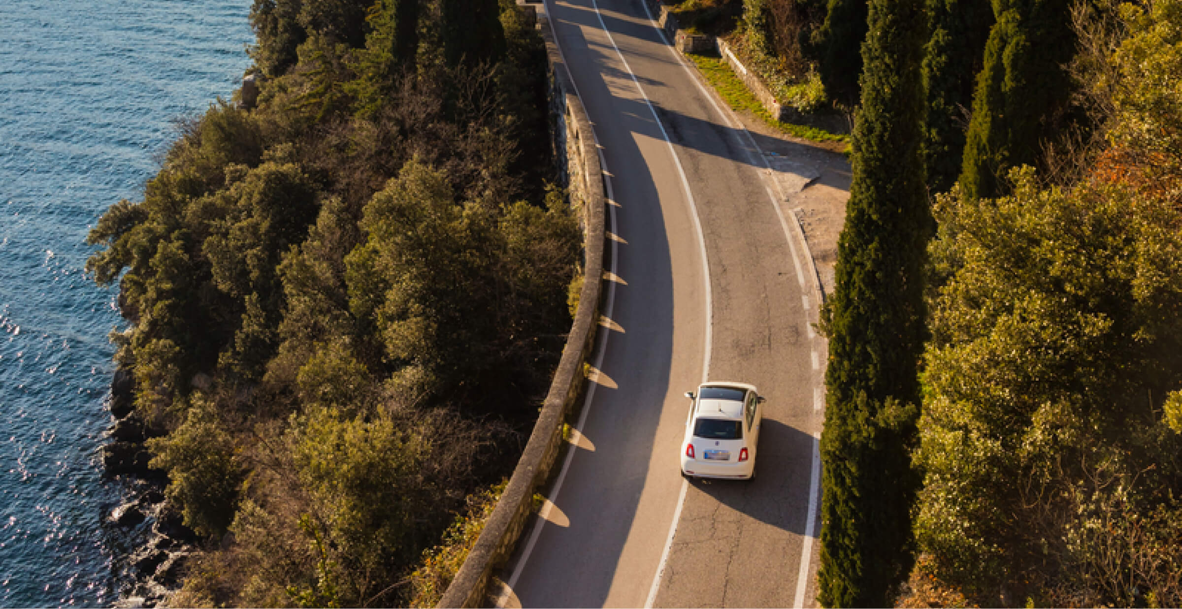 Aerial view of a white car driving on a winding coastal road. The road is surrounded by lush green trees on one side and a deep blue body of water on the other, offering a picturesque landscape. The scene evokes a peaceful and relaxing journey, with the vehicle navigating along the cliffside path under the clear sky.