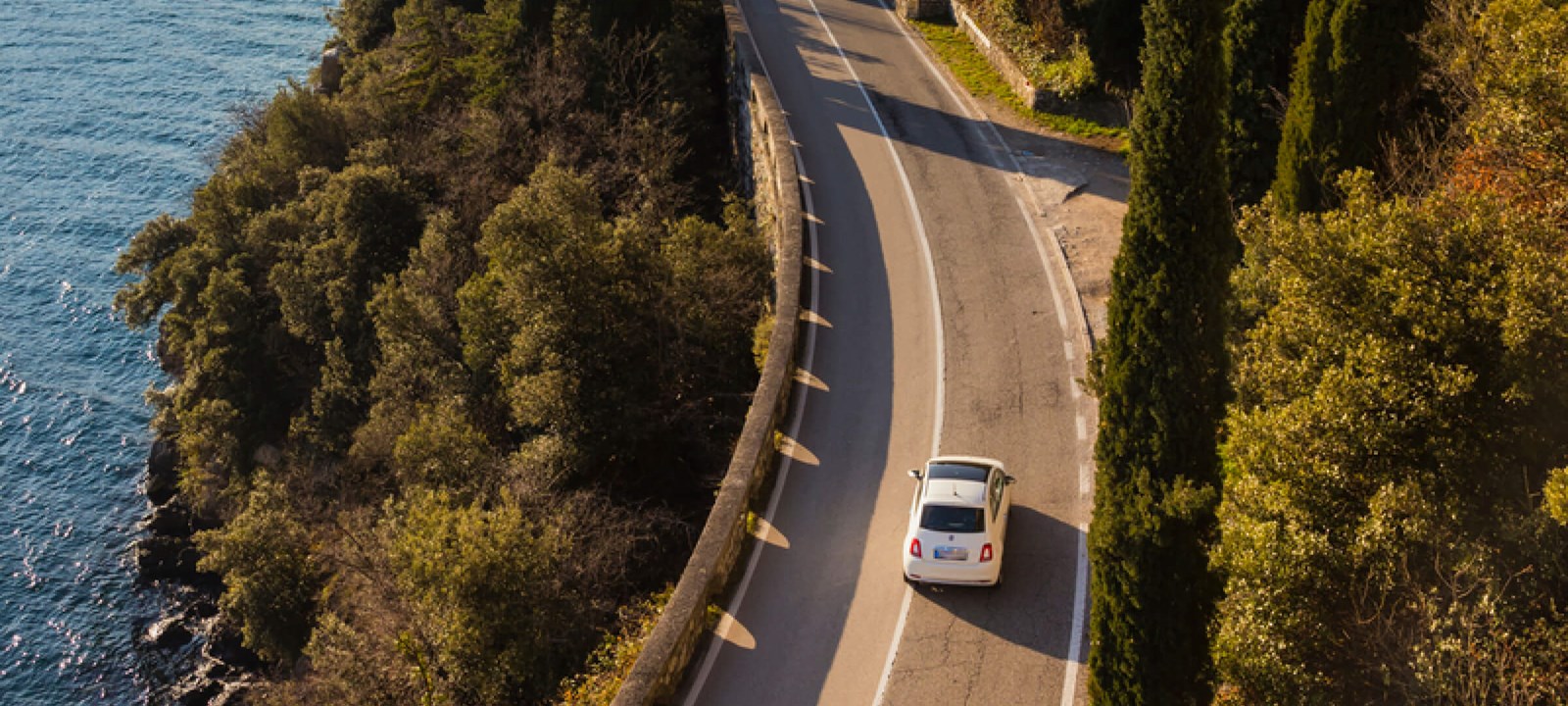 Aerial view of a white car driving on a winding coastal road. The road is surrounded by lush green trees on one side and a deep blue body of water on the other, offering a picturesque landscape. The scene evokes a peaceful and relaxing journey, with the vehicle navigating along the cliffside path under the clear sky.