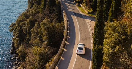Aerial view of a white car driving on a winding coastal road. The road is surrounded by lush green trees on one side and a deep blue body of water on the other, offering a picturesque landscape. The scene evokes a peaceful and relaxing journey, with the vehicle navigating along the cliffside path under the clear sky.