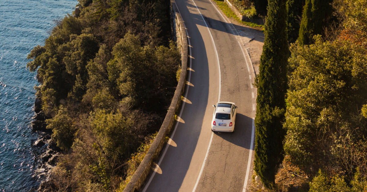 Aerial view of a white car driving on a winding coastal road. The road is surrounded by lush green trees on one side and a deep blue body of water on the other, offering a picturesque landscape. The scene evokes a peaceful and relaxing journey, with the vehicle navigating along the cliffside path under the clear sky.