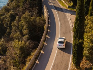 Aerial view of a white car driving on a winding coastal road. The road is surrounded by lush green trees on one side and a deep blue body of water on the other, offering a picturesque landscape. The scene evokes a peaceful and relaxing journey, with the vehicle navigating along the cliffside path under the clear sky.