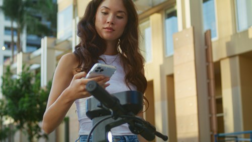 A young woman stands outdoors, focused on her smartphone while interacting with an electric scooter or bike, likely using an app to unlock or navigate it. She is dressed casually in a white tank top and jeans, with long, wavy hair. The background features a modern urban setting with beige buildings and greenery, suggesting a relaxed city environment. The image highlights the use of technology for convenient urban mobility.