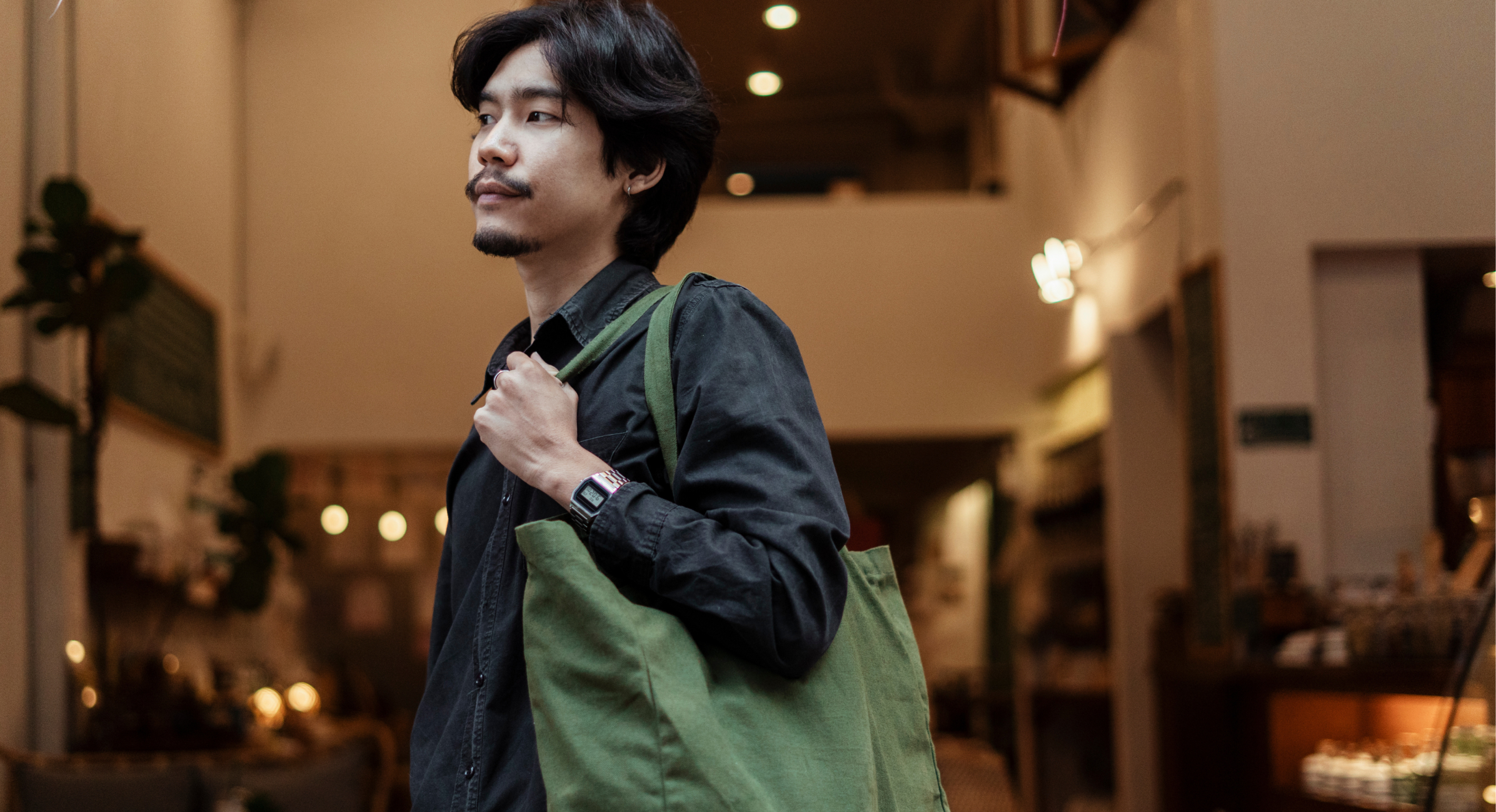 A man with shoulder-length hair, wearing a black shirt, holds a green reusable shopping bag over his shoulder while standing inside a well-lit store.