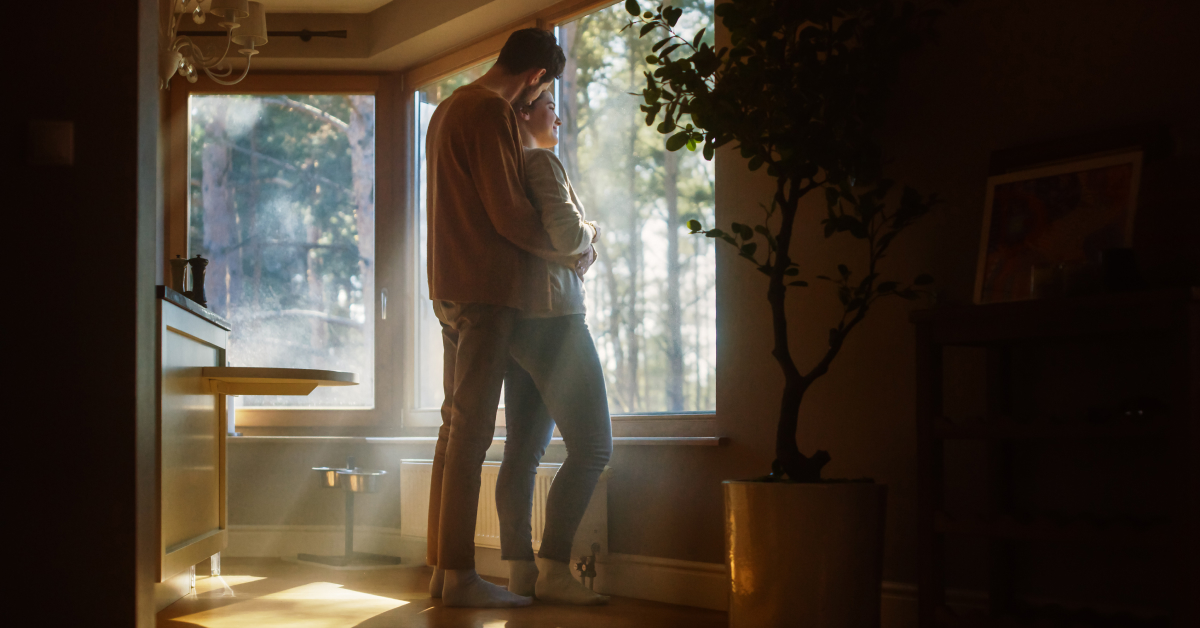 A couple stands together in a cozy living room, embracing and looking out a large window with sunlight streaming in. The room is softly lit, with warm tones and a peaceful atmosphere. A potted plant and framed artwork are visible in the background, adding to the serene setting. The couple appears to be enjoying a quiet moment together, appreciating the view of the trees outside.