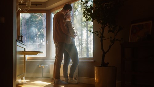 A couple stands together in a cozy living room, embracing and looking out a large window with sunlight streaming in. The room is softly lit, with warm tones and a peaceful atmosphere. A potted plant and framed artwork are visible in the background, adding to the serene setting. The couple appears to be enjoying a quiet moment together, appreciating the view of the trees outside.