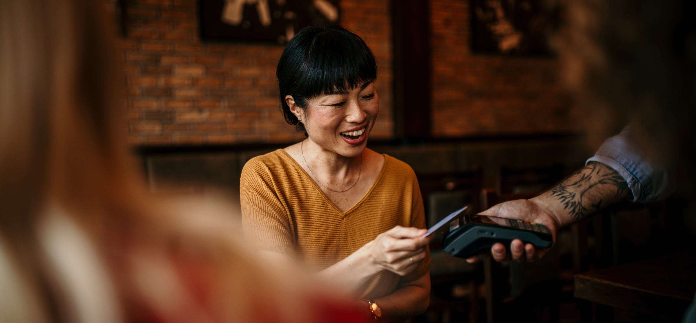 A woman in a mustard-colored top smiles as she pays using her card in a restaurant. She holds the card up to a payment terminal, while a tattooed server holds the machine. The background shows a cozy, brick-walled dining space with dim lighting, creating a warm and relaxed atmosphere.