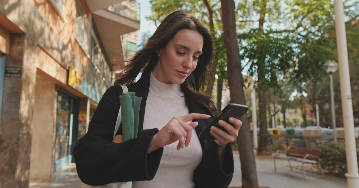 A woman carrying groceries walks outdoors while using her smartphone, surrounded by trees and buildings.