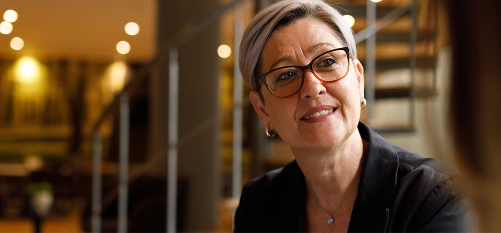 A woman with short gray hair and glasses is engaged in a conversation, smiling warmly. She is dressed professionally, and the background features a modern, softly-lit office environment with a staircase and blurred lighting.