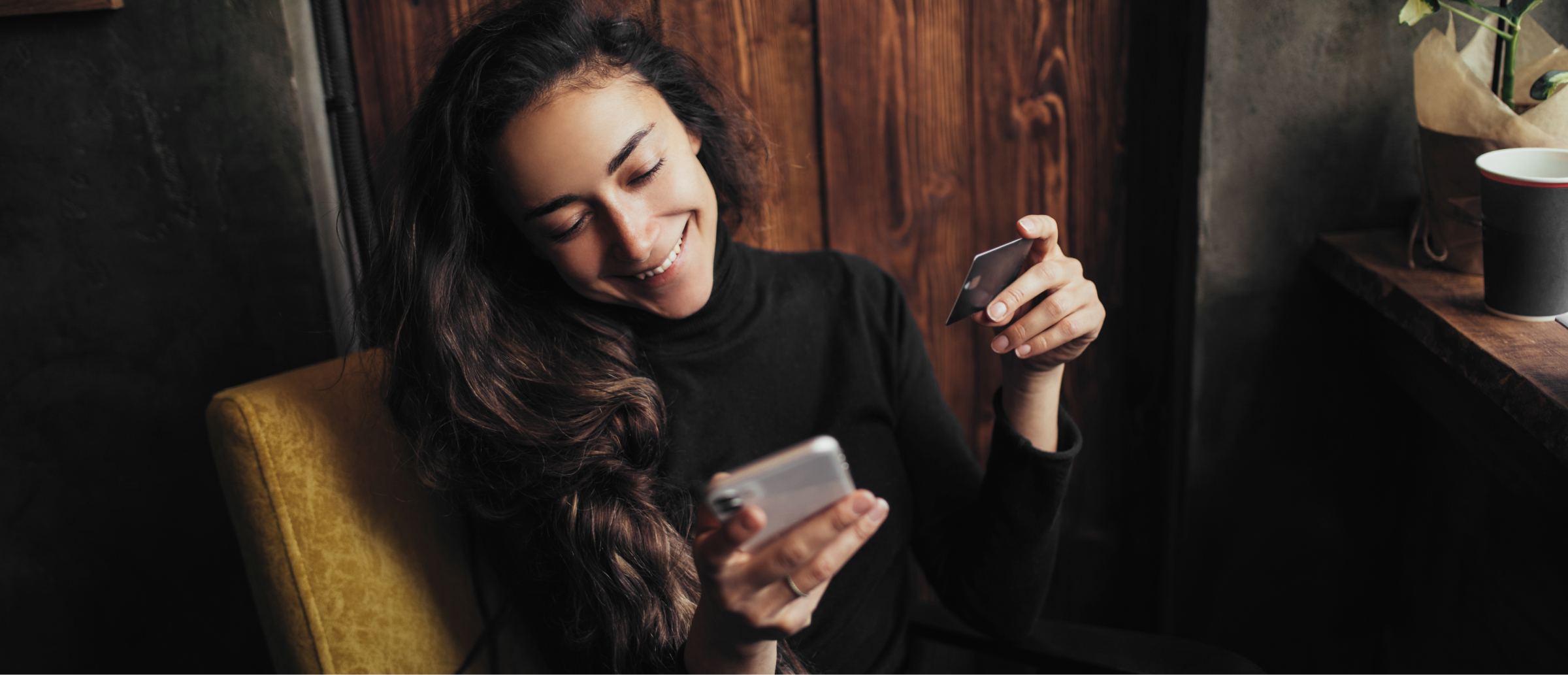 A woman with long dark hair smiles as she activates her bank card on her mobile phone. She is seated in a cozy space, holding the card in one hand and her phone in the other, enjoying a peaceful moment.