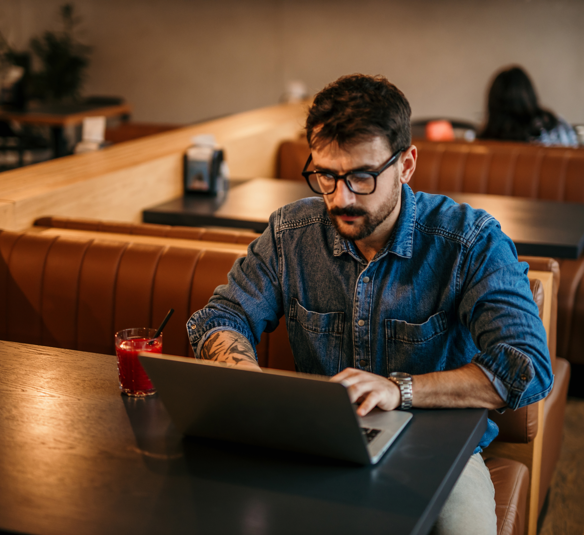 A man wearing glasses and a denim shirt works on his laptop in a café. He is focused on the screen, with a glass of juice next to him on the table. The cozy café atmosphere suggests a productive and relaxed workspace.