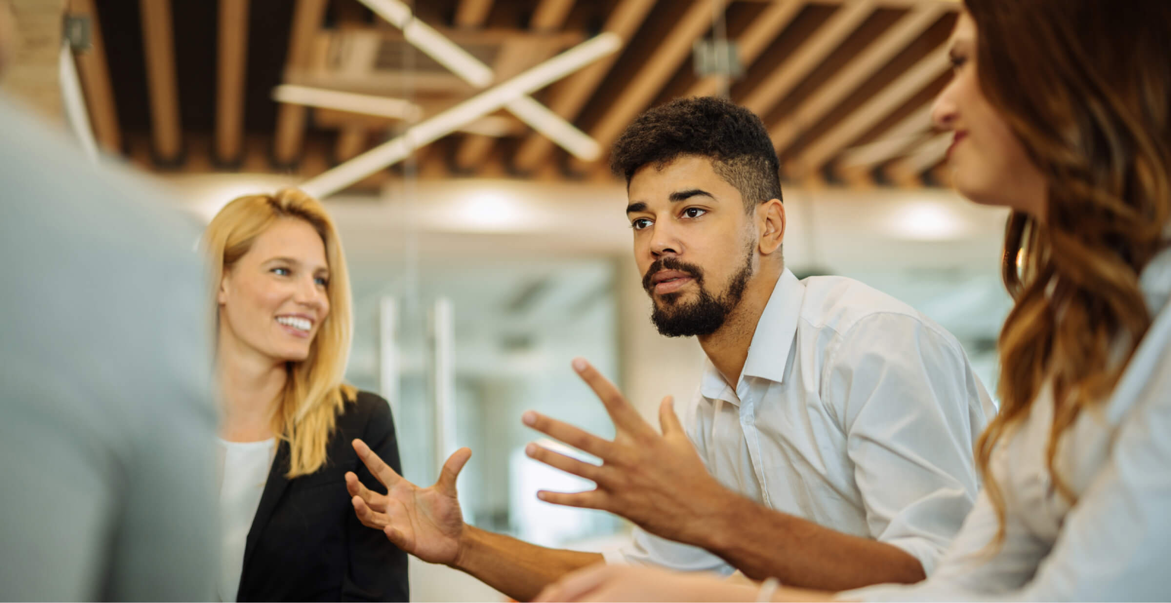 A group of colleagues in a modern office engaged in a discussion. A man with curly hair and a beard gestures with his hands while speaking, and two women, one with blonde hair and the other with brown, listen attentively. The background features a ceiling with wooden beams and contemporary lighting, creating a collaborative and dynamic atmosphere. The team appears focused on exchanging ideas, likely about AI