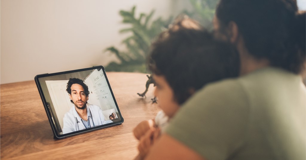A mother and child are sitting together at a table, engaged in a video call with a doctor on a tablet. The doctor, who appears to be wearing a white coat and has a stethoscope around his neck, is speaking to them from his office, with a whiteboard visible in the background. The setting is calm and homey, with a small toy dinosaur on the table near the child. The image highlights the convenience and comfort of telemedicine, allowing patients to consult with healthcare professionals from their own home