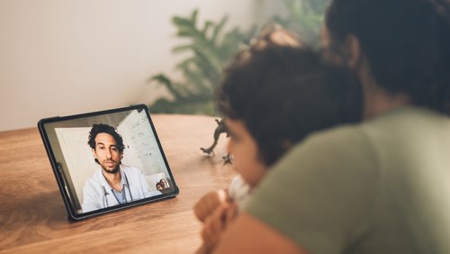 A mother and child are sitting together at a table, engaged in a video call with a doctor on a tablet. The doctor, who appears to be wearing a white coat and has a stethoscope around his neck, is speaking to them from his office, with a whiteboard visible in the background. The setting is calm and homey, with a small toy dinosaur on the table near the child. The image highlights the convenience and comfort of telemedicine, allowing patients to consult with healthcare professionals from their own home