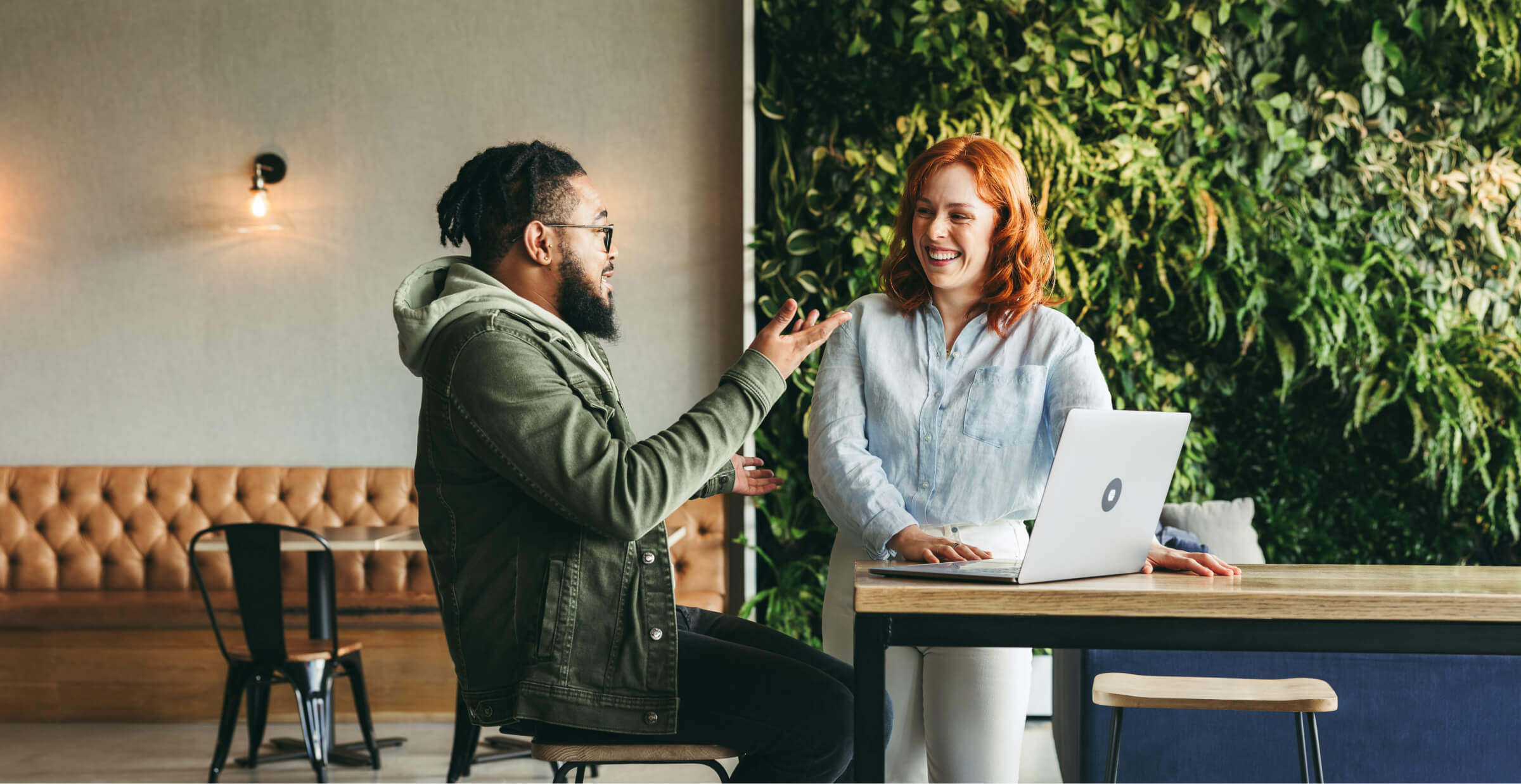A man and woman are engaged in a lively discussion in a modern, casual workspace. The man, wearing a green jacket, gestures with his hands while seated, and the woman, with red hair, stands next to him, smiling with a laptop on the table. The background features a leather bench, modern lighting, and a wall covered in green plants, creating a warm and relaxed atmosphere. The scene conveys collaboration and friendly communication.