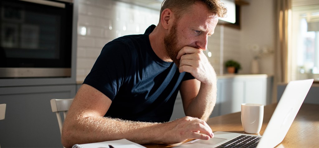 A man is sitting at a kitchen table, deeply focused on his laptop. He has a thoughtful expression, with one hand resting on his chin and the other on the keyboard. The setting is a modern kitchen with soft lighting, creating a calm atmosphere. A white coffee mug sits on the table beside him, and a notebook with a pen is visible in the corner. The image captures a moment of concentration, likely working from home or doing research online.