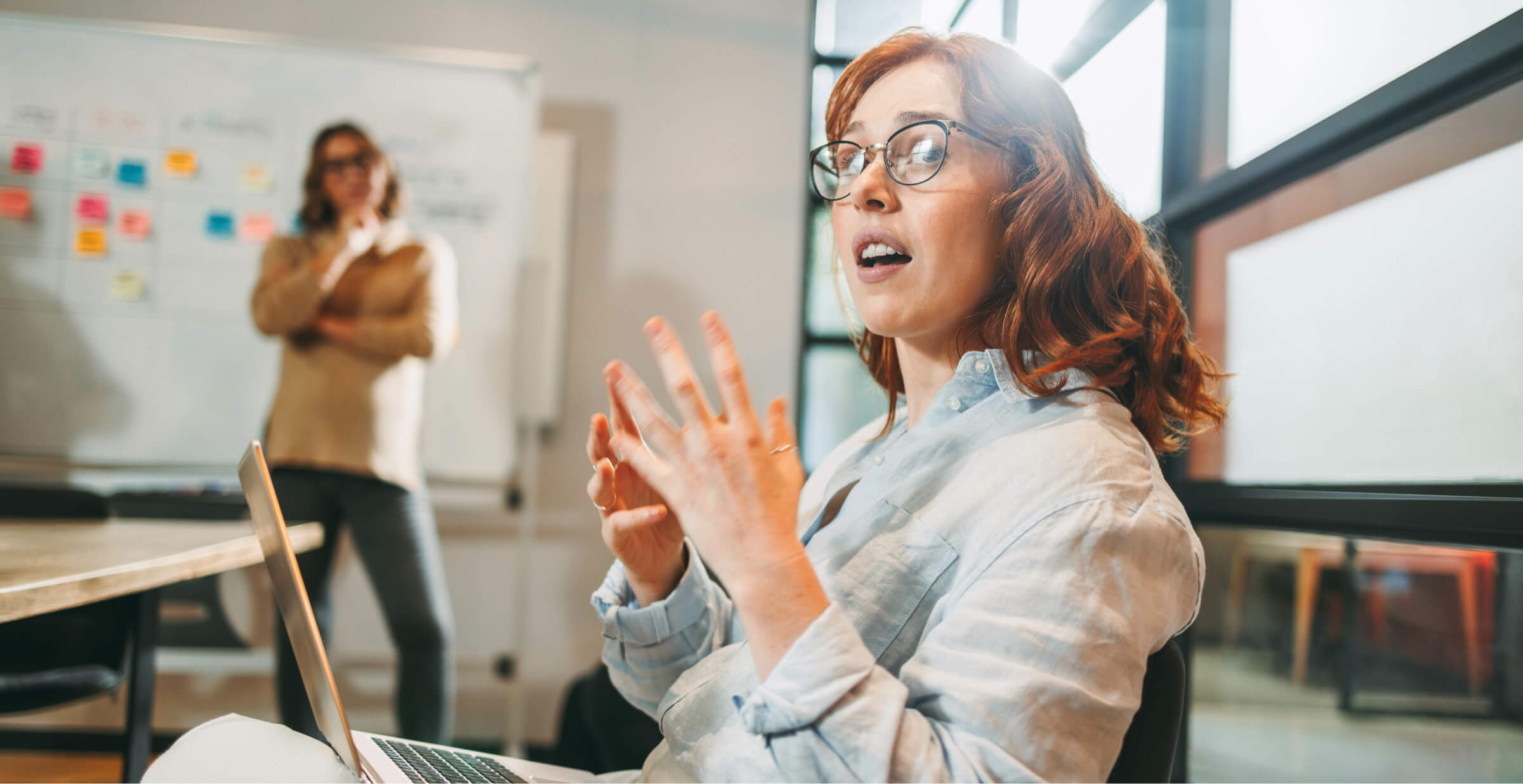 A woman with red hair and glasses gestures as she speaks during a meeting, with a laptop open on her lap. In the background, another team member stands by a whiteboard covered with colorful sticky notes, observing the discussion. The office environment is modern and collaborative, with large windows letting in natural light. The scene captures a creative brainstorming session with engaged participants.