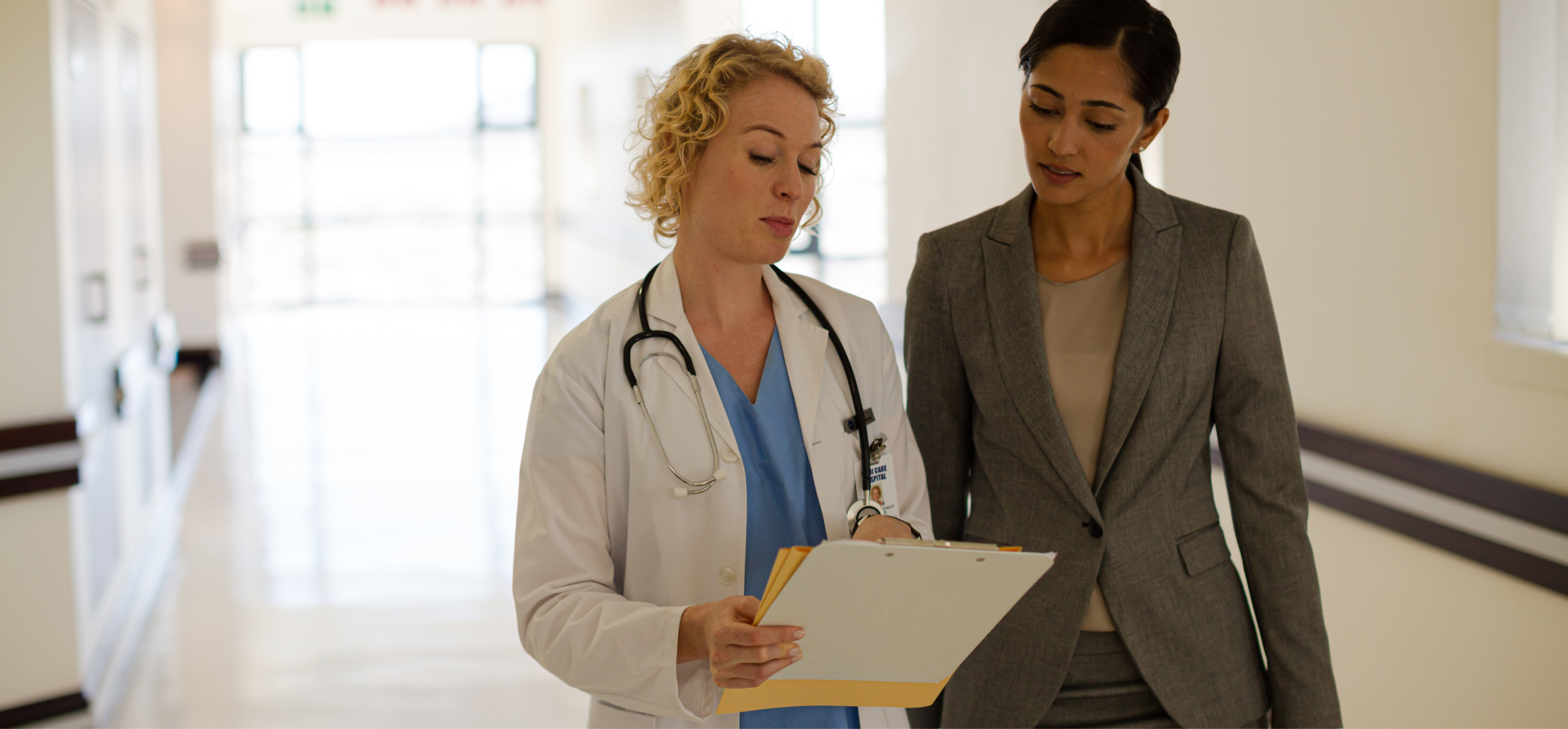 A female doctor and a professional woman are walking together down a brightly lit hospital hallway. The doctor, wearing a white coat and stethoscope, is holding a clipboard and discussing its contents with the woman, who is dressed in a gray business suit. The two appear to be collaborating, possibly reviewing medical documents or a patient case. The background features clean, modern hospital decor, emphasizing a professional and clinical environment. The image highlights a moment of teamwork and communication in a healthcare setting.