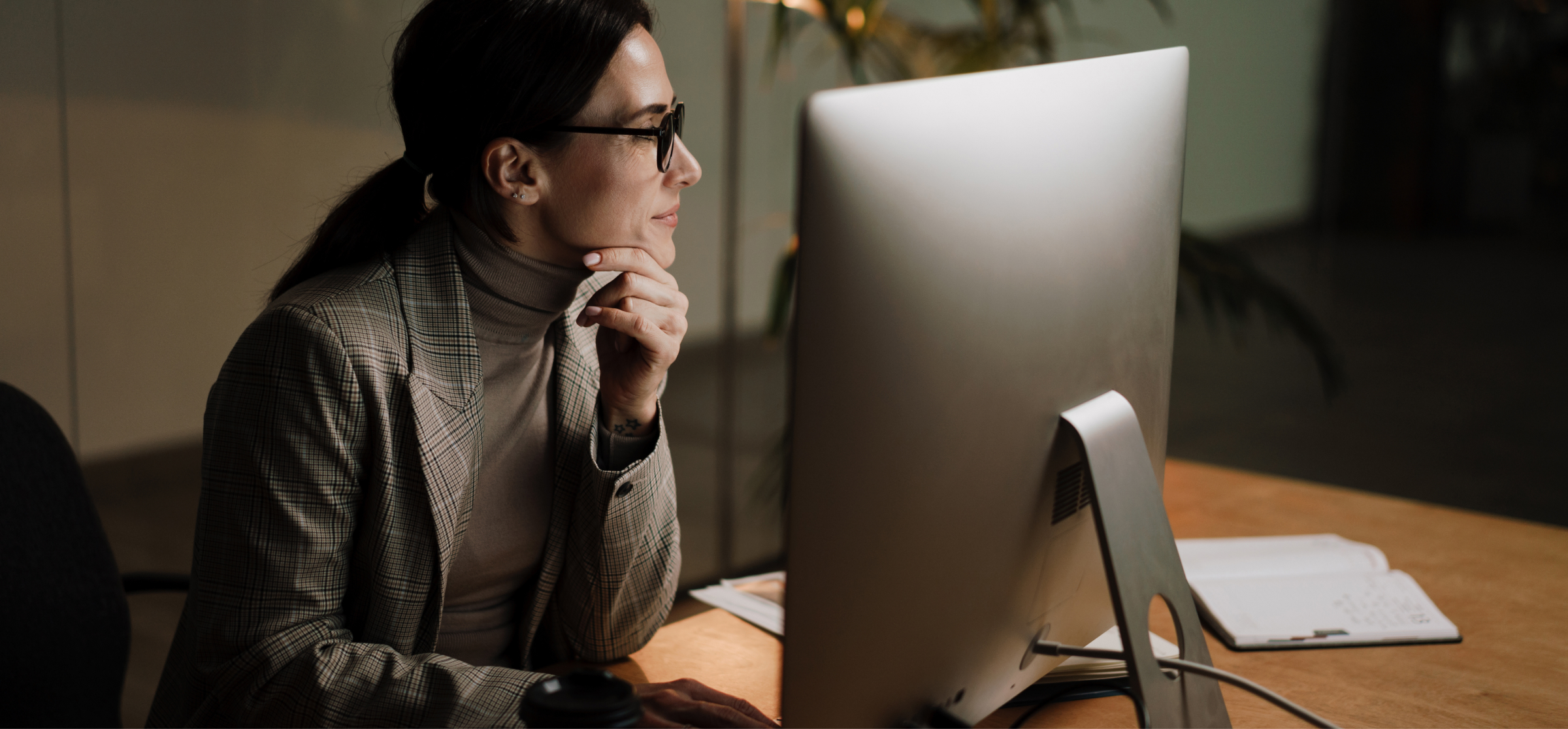 A woman wearing glasses and a checkered blazer is sitting at a desk, deeply focused on her work on a desktop computer. She rests her chin on her hand as she concentrates on the screen. The room is dimly lit, with soft lighting creating a calm and professional atmosphere. A notebook with handwritten notes is open on the desk next to her, indicating that she might be taking notes or referring to information. The image captures a moment of focused productivity in a work setting.