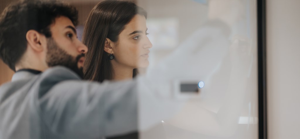 A man and a woman are working together, focusing on a whiteboard or screen in a collaborative environment. The man is pointing at something on the board while the woman observes intently. Both are dressed in professional attire, with the woman wearing earrings and having long, dark hair. The image captures a moment of concentration and teamwork, with both individuals engaged in problem-solving or strategizing. The scene emphasizes collaboration and attention to detail in a professional setting.