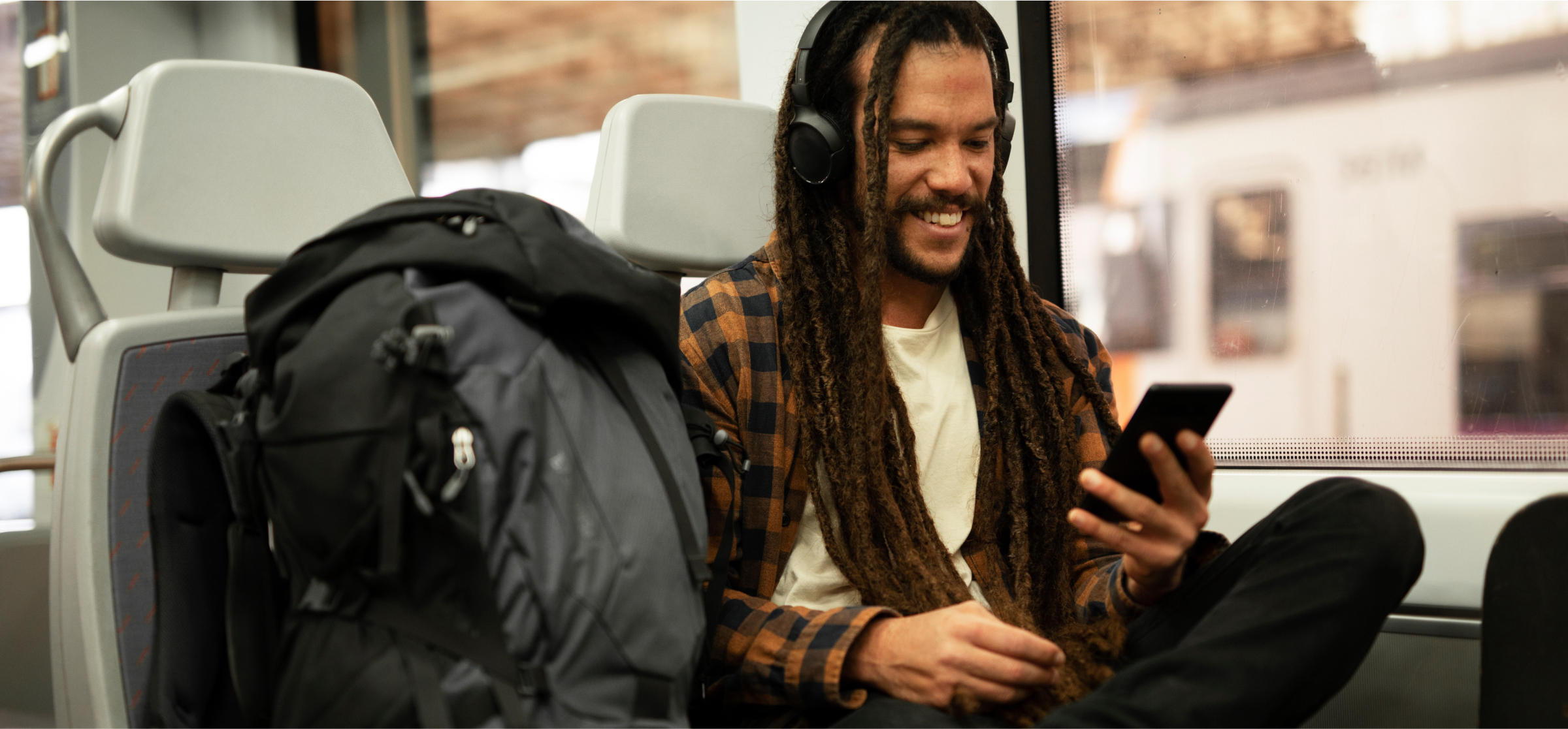  A man with long dreadlocks is sitting on a train, smiling as he looks at his smartphone. He is wearing headphones and a casual plaid shirt, enjoying content on his phone during the ride. Next to him is a large backpack placed on the seat. The background shows a train station platform through the window, with the train in motion. The image conveys a relaxed and pleasant travel experience, with technology and entertainment making the journey enjoyable.