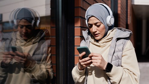 A woman wearing a hijab, headphones, and a puffy vest, standing against a brick wall outdoors, looking at her smartphone while her reflection appears on the glass next to her.