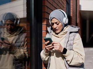 A woman wearing a hijab, headphones, and a puffy vest, standing against a brick wall outdoors, looking at her smartphone while her reflection appears on the glass next to her.