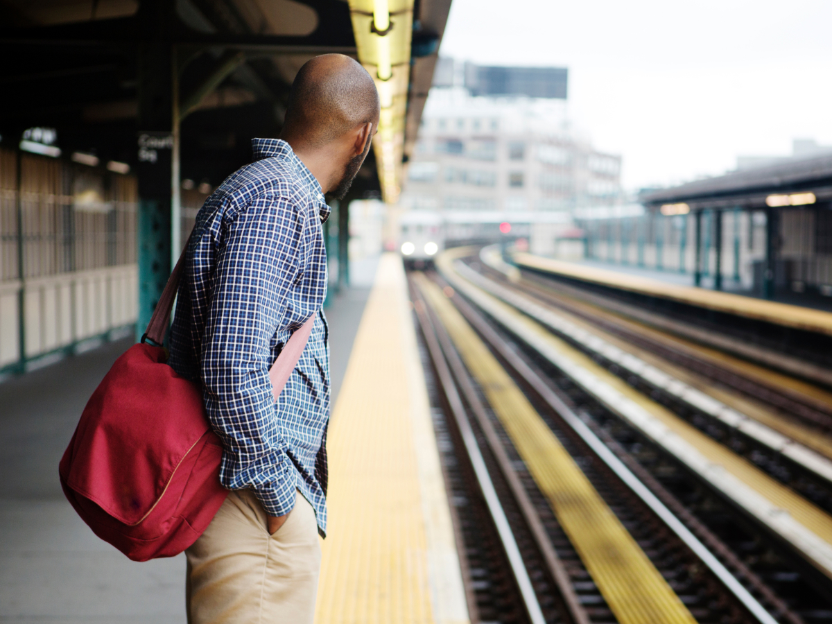 A man wearing a checkered shirt and carrying a red bag stands on a train platform, looking down the tracks as a train approaches in the distance.