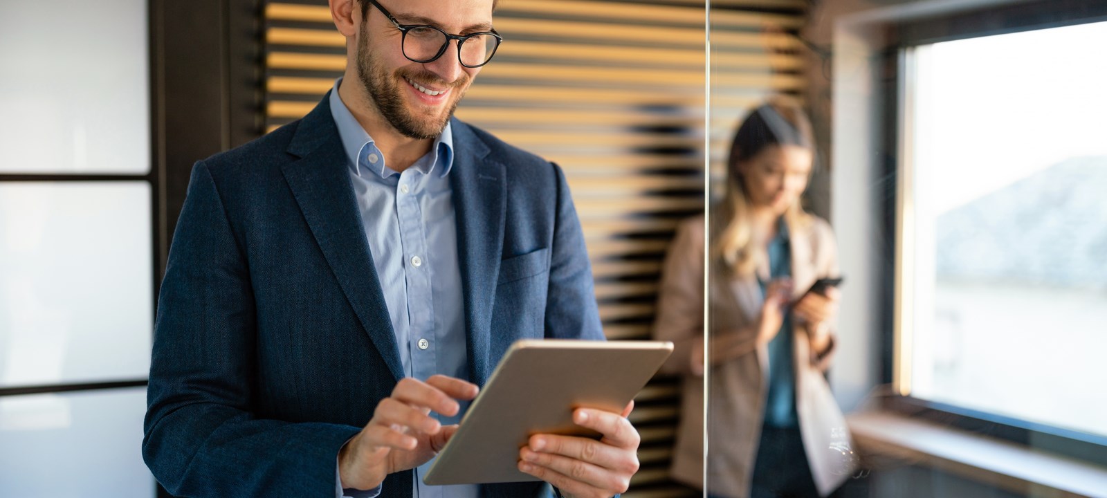 A man in a business suit and glasses is smiling while working on a tablet in a modern office. He is standing near a glass wall, with a woman in the background using her phone. The man appears focused and engaged, likely reviewing or analyzing something on the tablet. The setting is professional, with warm lighting and a sleek design, featuring wooden panels on the wall. The image conveys a sense of productivity, technology use, and a collaborative work environment.