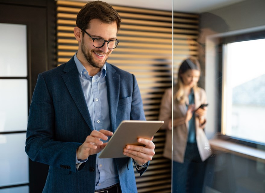 A man in a business suit and glasses is smiling while working on a tablet in a modern office. He is standing near a glass wall, with a woman in the background using her phone. The man appears focused and engaged, likely reviewing or analyzing something on the tablet. The setting is professional, with warm lighting and a sleek design, featuring wooden panels on the wall. The image conveys a sense of productivity, technology use, and a collaborative work environment.