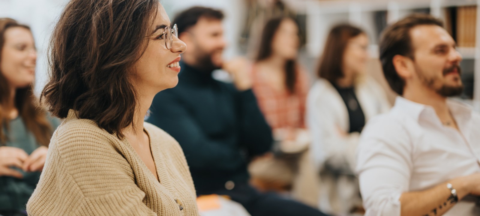 A group of people is sitting in a classroom or conference setting, engaged and smiling as they listen to a speaker. The focus is on a woman in the foreground, who has shoulder-length brown hair and is wearing glasses and a beige cardigan.