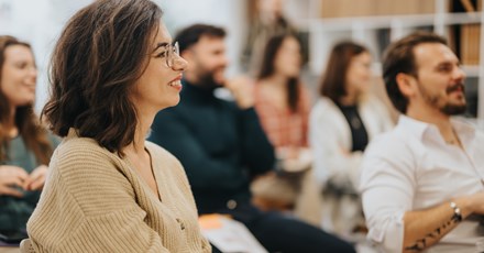 A group of people is sitting in a classroom or conference setting, engaged and smiling as they listen to a speaker. The focus is on a woman in the foreground, who has shoulder-length brown hair and is wearing glasses and a beige cardigan.