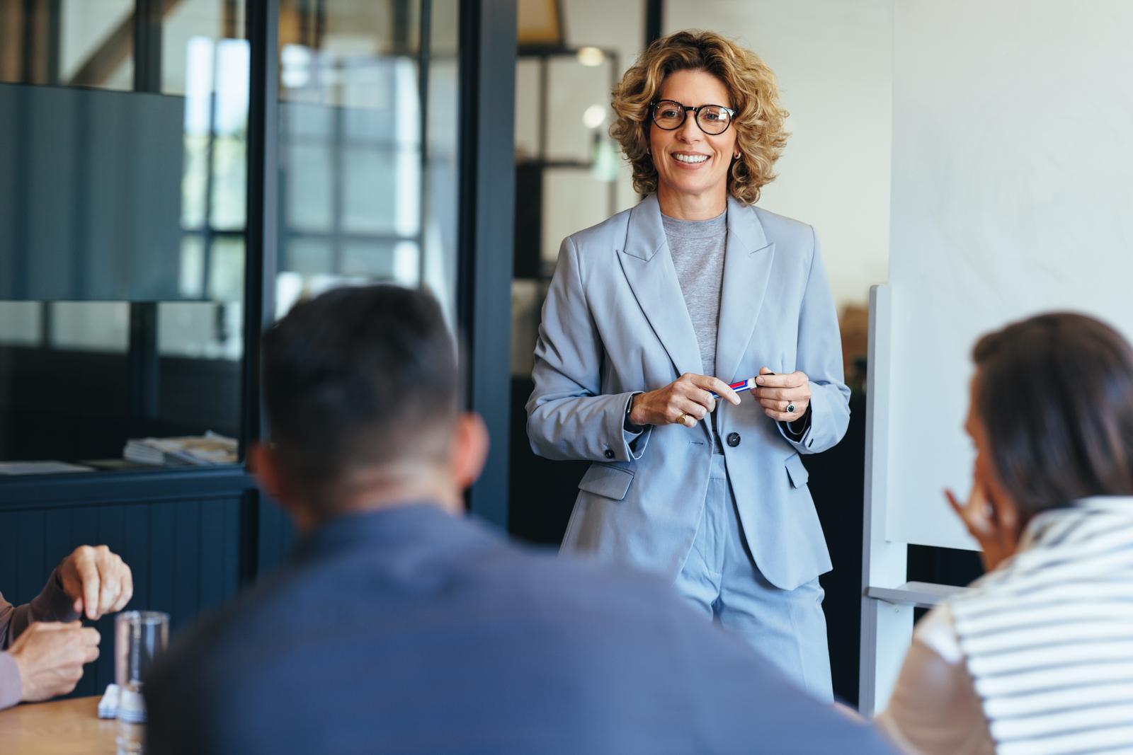 A businesswoman in a light gray suit smiles while leading a discussion with her team in a modern office setting.