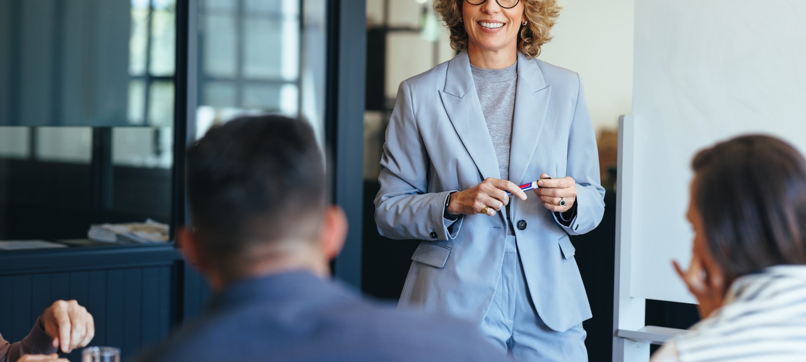 A businesswoman in a light gray suit smiles while leading a discussion with her team in a modern office setting.