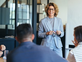 A businesswoman in a light gray suit smiles while leading a discussion with her team in a modern office setting.